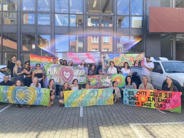 Volontäre aus verschiedenen Ländern erstellen Friedensbanner bei einer Fortbildung im Kinder- und Jugendateier Roter Hahn her. Auf dem Foto zeigen sie in einem Gruppenbild die mehrsprachigen Banner