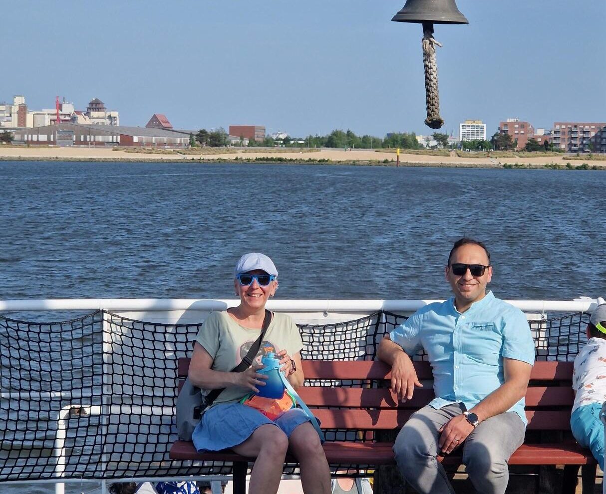 Zwei Personen mit Sonnenbrillen sitzen auf einer Bank auf einer Fähre. Im Hintergrund sind Wasser und ein Sandstrand zu sehen.