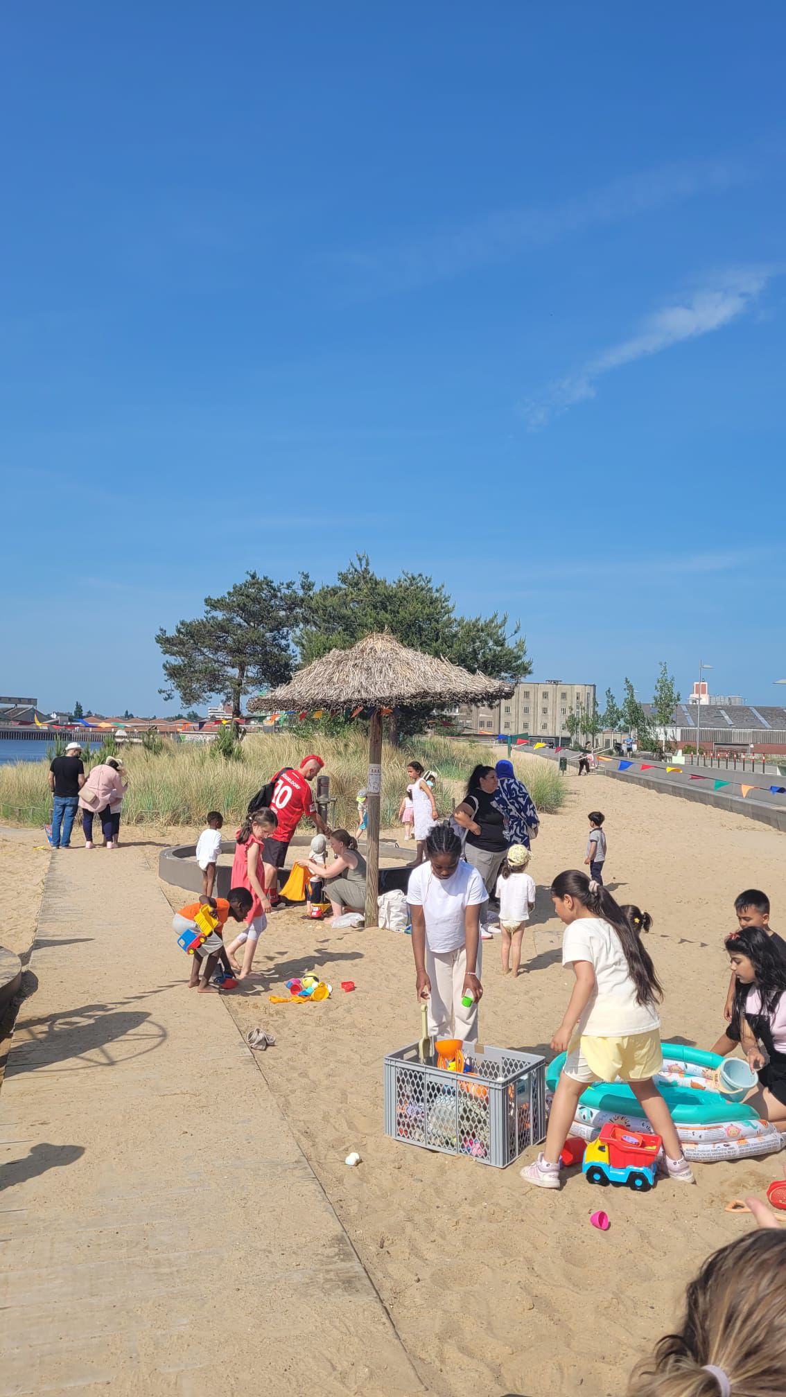 An einem Sandstrand sind Bänke mit Erwachsenen und Kindern zu sehen. Im hintergrund gibt es Wasser.