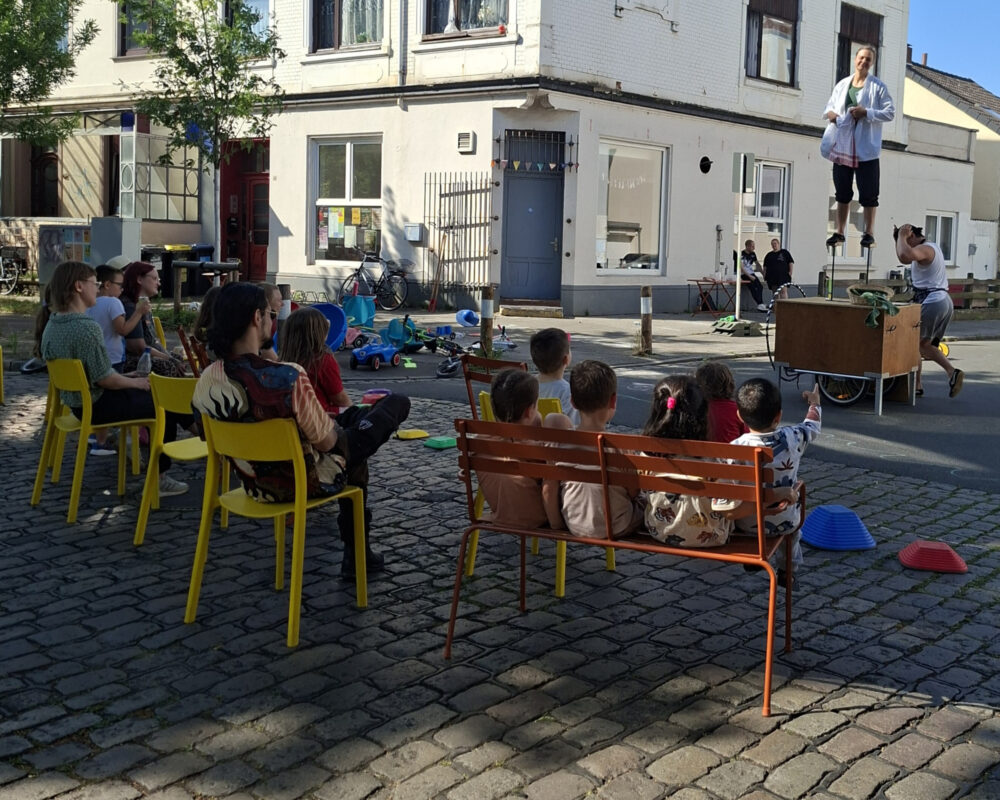 Auf einer Straße sitzen Menschen auf Stühlen und schauen einer Akrobatik Show zu