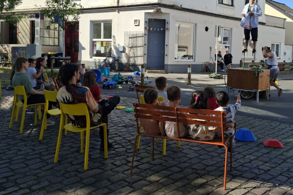 Auf einer Straße sitzen Menschen auf Stühlen und schauen einer Akrobatik Show zu
