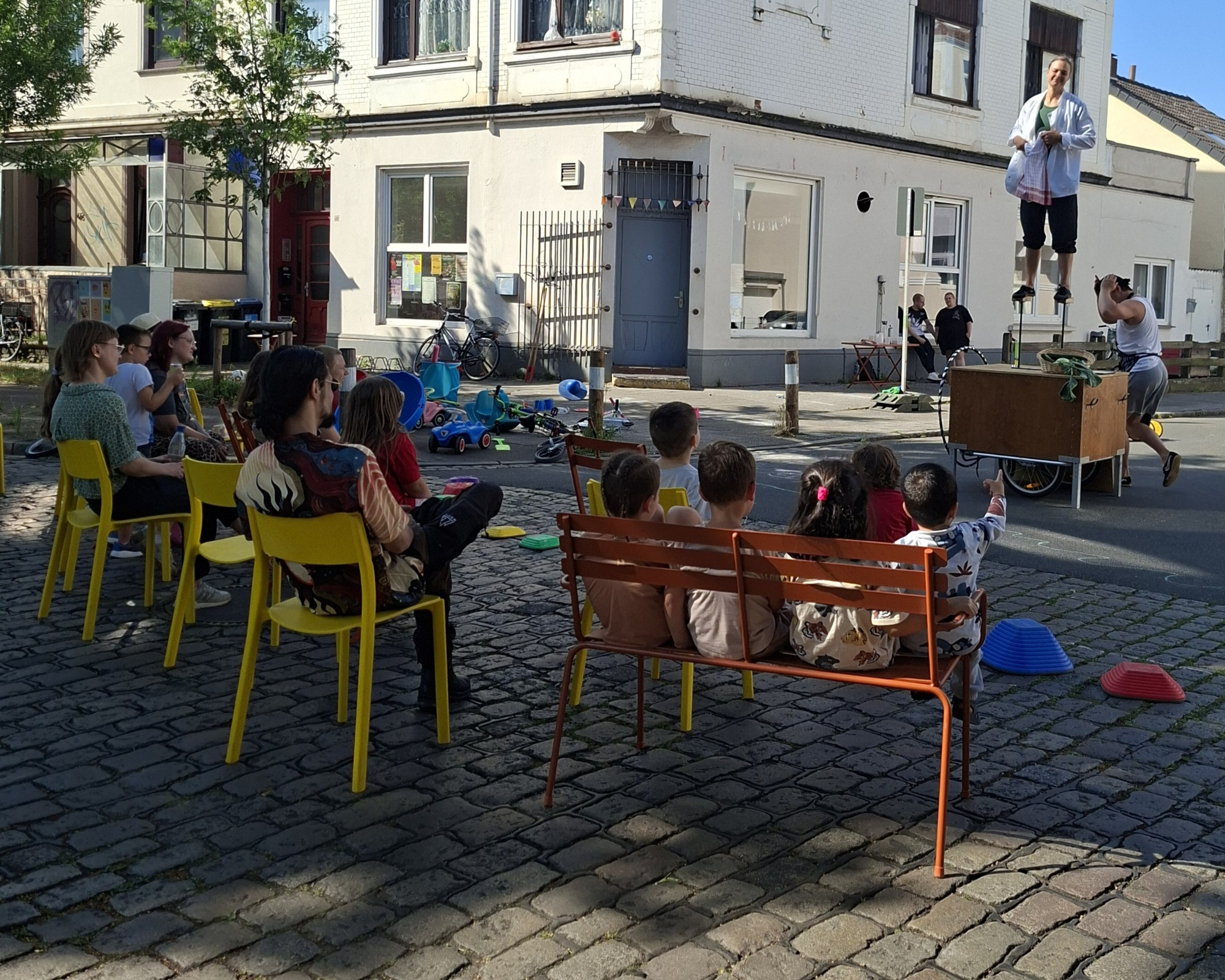 Auf einer Straße sitzen Menschen auf Stühlen und schauen einer Akrobatik Show zu