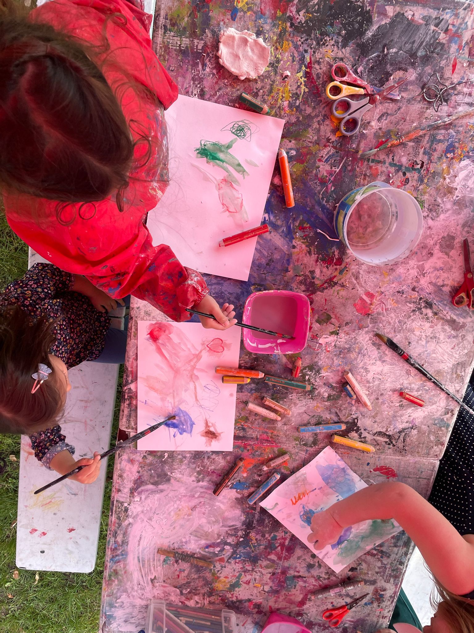 Aus der Vogelperspektive wird auf einen Arbeitstisch geschaut an dem zwei Kinder ihre Pinsel in Wasser tauchen. Auf dem Tisch liegt Papier, auf dem Malereien zu sehen sind. Der Tisch ist voller Farbkleckse.