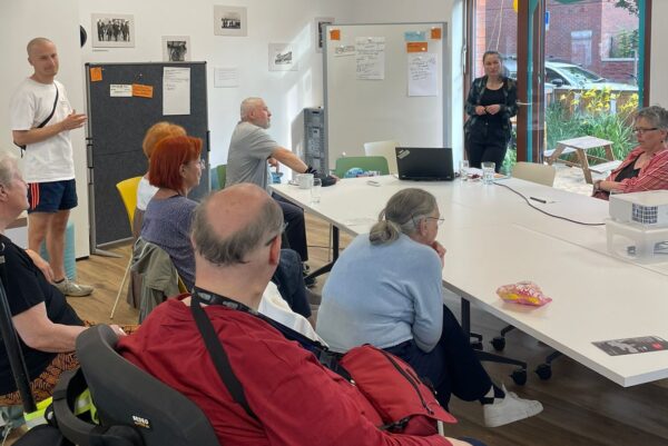 In einem Raum sitzen Menschen rund um einen Tisch. Zwei Personen stehen. Auf dem Tisch steht ein Beamer.