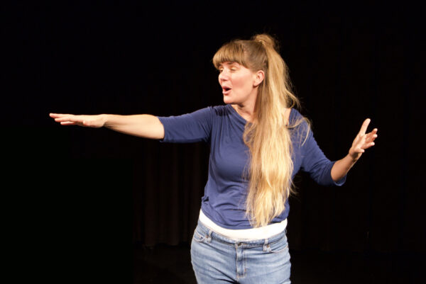 Vor einer schwarzen Leinwand befindet sich mit ausdruckstarker Gestik und mimik eine darstellende Künstlerin.