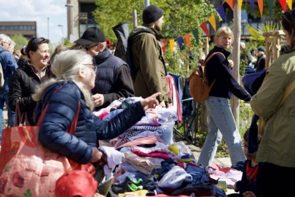 Bunte Wimpelgirlanden hängen über mehreren Tischen, die mit Kleidung und Textilien bedeckt sind. Menschen stehen dicht beieinander, betrachten die ausgelegten Sachen und greifen nach einzelnen Kleidungsstücken. Im Hintergrund sind moderne Gebäude mit großen Fenstern und Balkonen sowie grüne Bäume zu sehen.