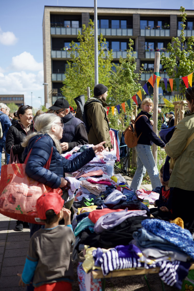 Bunte Wimpelgirlanden hängen über mehreren Tischen, die mit Kleidung und Textilien bedeckt sind. Menschen stehen dicht beieinander, betrachten die ausgelegten Sachen und greifen nach einzelnen Kleidungsstücken. Im Hintergrund sind moderne Gebäude mit großen Fenstern und Balkonen sowie grüne Bäume zu sehen.