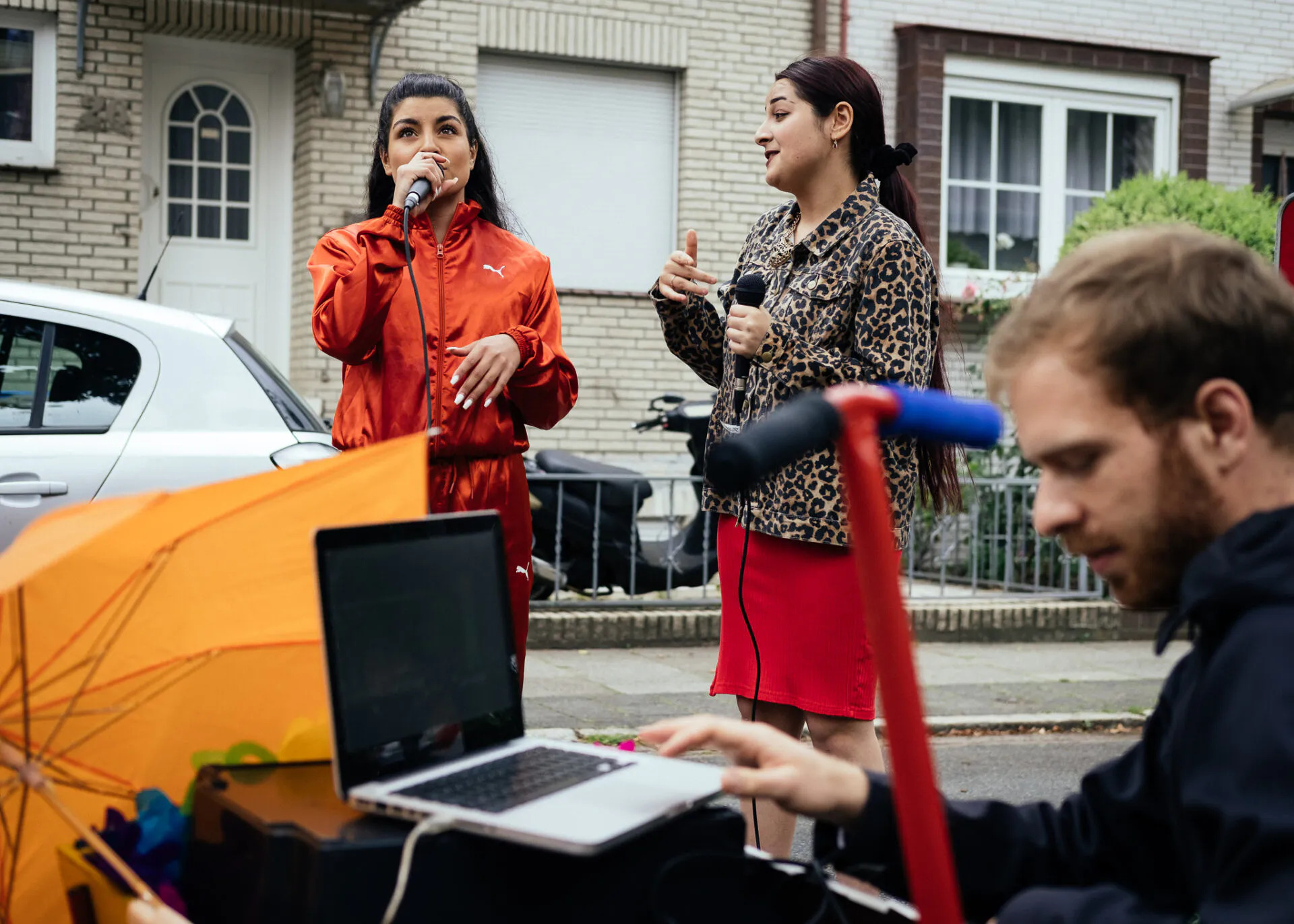 Zwei Sängerinnen rappen bei einem Straßenfest. Rechts vor ihnen sitzt eine Person am Mischpult und Laptop. Ein Regenschirm ist links davon gespannt.