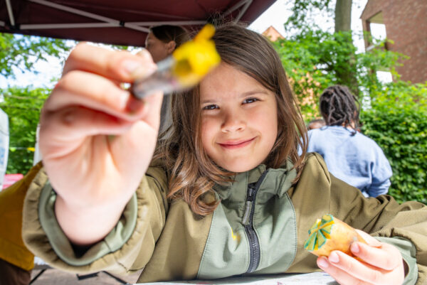 Ein Kind hält einen Pinsel mit gelber Farbe in der einen Hand richtung Kamera und lächelt. in der anderen Hand hält sie eine Kartoffel, die für einen Kartoffeldruck zugeschnitten ist.