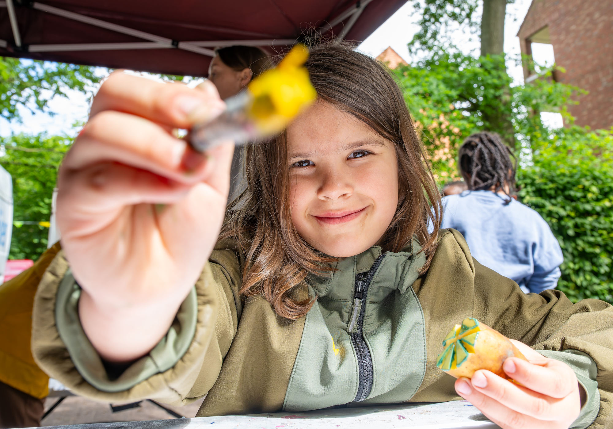 Ein Kind hält einen Pinsel mit gelber Farbe in der einen Hand richtung Kamera und lächelt. in der anderen Hand hält sie eine Kartoffel, die für einen Kartoffeldruck zugeschnitten ist.