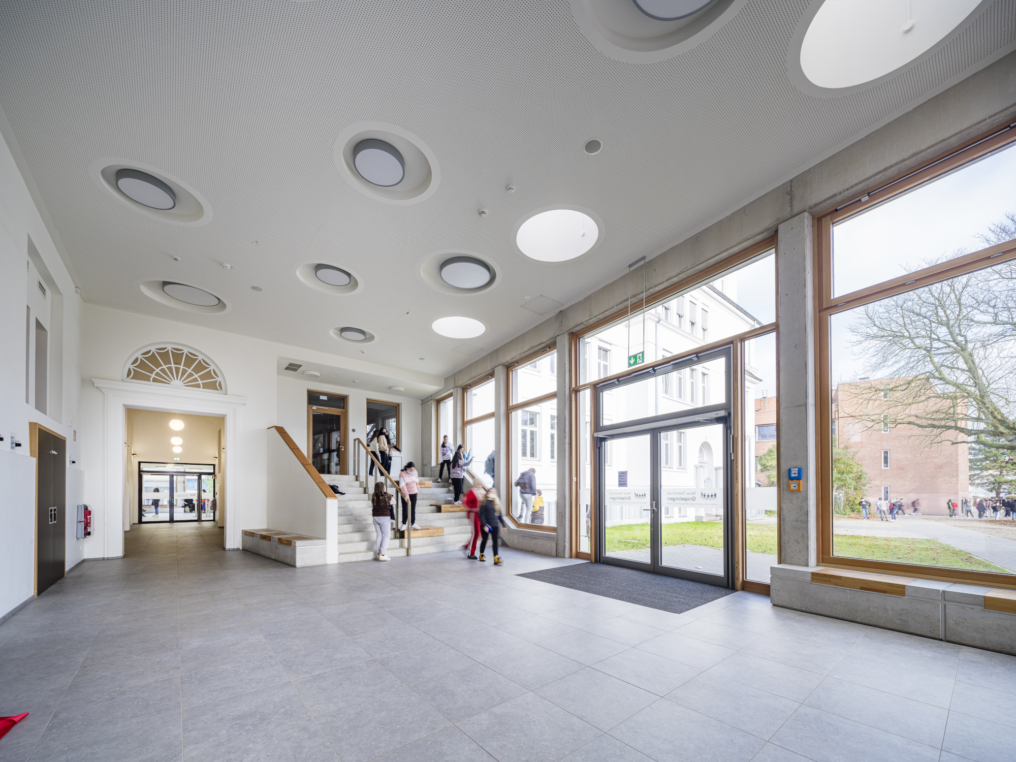 Ein lichtdurchflutetes Foyer einer Schule ist zu sehen. Durch die Fensterscheiben sind weitere Gebäude zun ein Baum zu sehen. Kinder laufen eine Treppe hinunter.