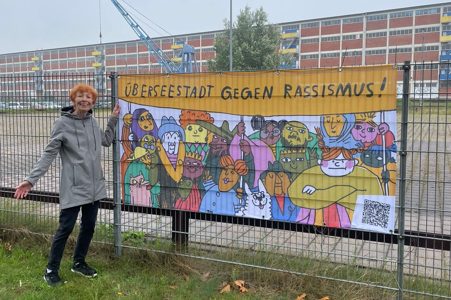 Ein Banner mit der Aufschrift "Überseestadt gegen Rassismus" an einem Zaun. Eine Frau steht links daneben und schaut in die Kamera.