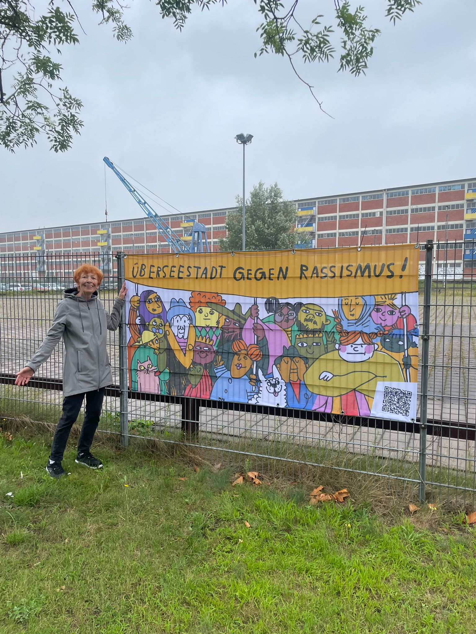 Ein Banner mit der Aufschrift "Überseestadt gegen Rassismus" an einem Zaun. Eine Frau steht links daneben und schaut in die Kamera.