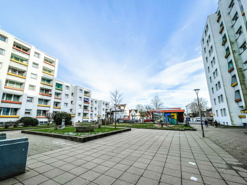 Zwischen zwei großen Hochhäusern ist ein gepflasterter Platz mit Spielplatz zu sehen.