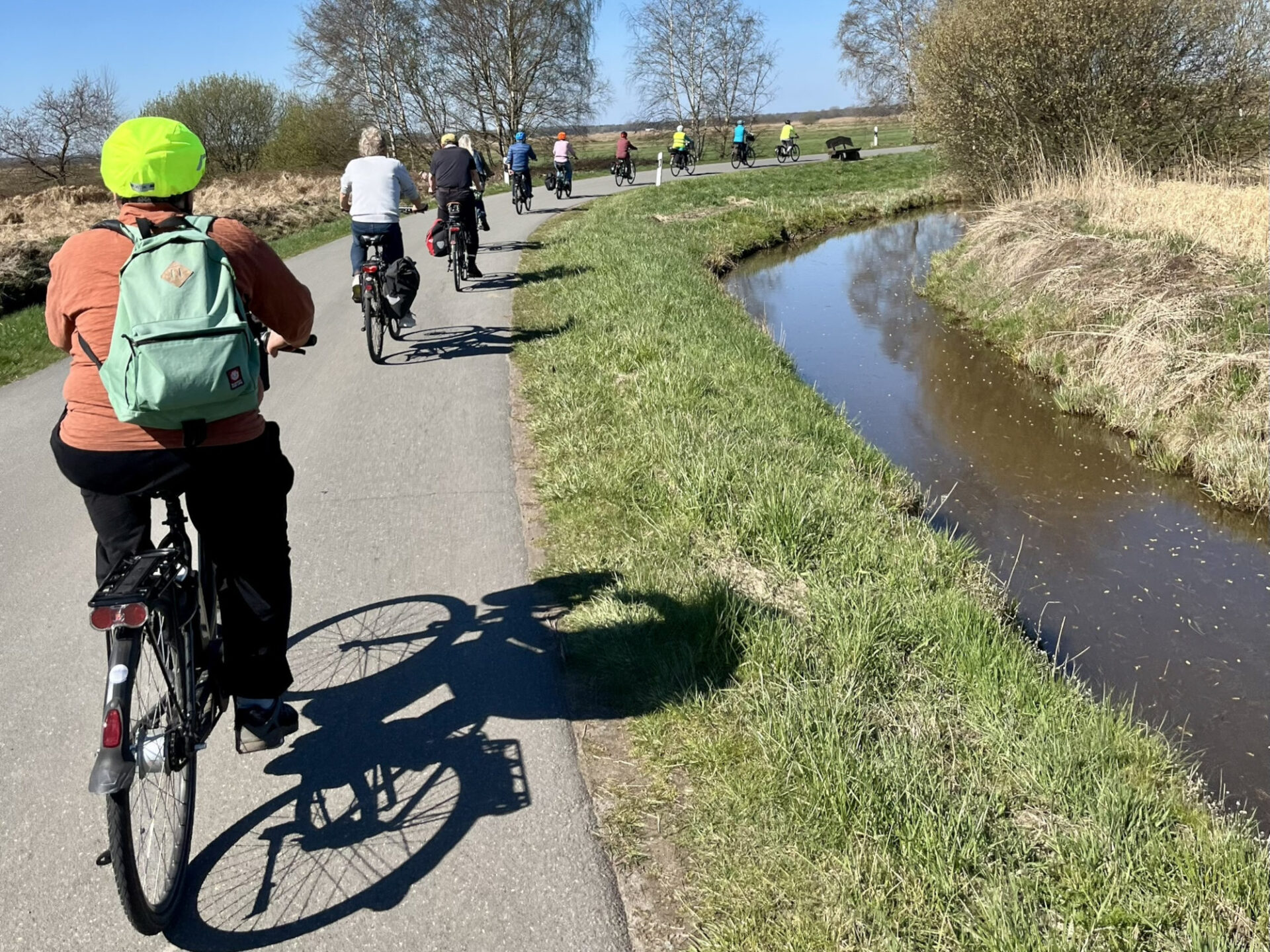 Auf einer asphaltierten Straße fahren Menschen Fahrrad. Rechts neben ihnen verläuft ein Kanal. Im Hintergrund sind Bäume zu sehen. Die Personen auf den Rädern sind von hinten zu sehen.