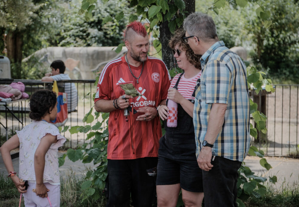 Drei erwachsene Personen stehen dicht beieinander ins Gespräch vertielt. Links daneben steht eine jugendliche Person mit Zopf. Eine Person hat einen roten Iro als Haarschnitt und trägt das Trickotoberteil von Fortuna Düsseldorf. Im Hintergrund befindet sich ein Spielplatz mit Bäumen.