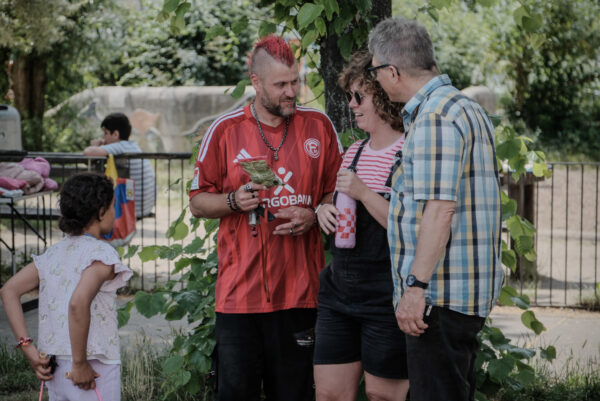 Drei erwachsene Personen stehen dicht beieinander ins Gespräch vertielt. Links daneben steht eine jugendliche Person mit Zopf. Eine Person hat einen roten Iro als Haarschnitt und trägt das Trickotoberteil von Fortuna Düsseldorf. Im Hintergrund befindet sich ein Spielplatz mit Bäumen.