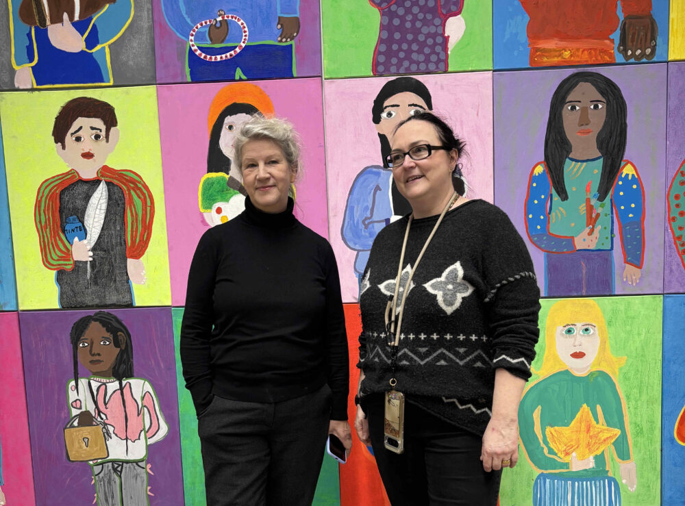 Voer einer Ausstellungswand mit den gemalten Porträts von Jugendlichen stehen zwei Personen und lächeln.