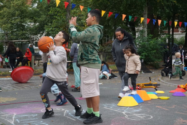 Bei einer Spielstrassenaktion befinden sich Kinder, Jugendliche und eine Erwachsene auf der Straße. Zwei Jugendliche haben Bälle in den Händen in Wurfhaltung. Die Erwachsene Person hilft einem Kinde beim Balancieren. Die Straße ist mit bunten Kreidefarben bemalt.