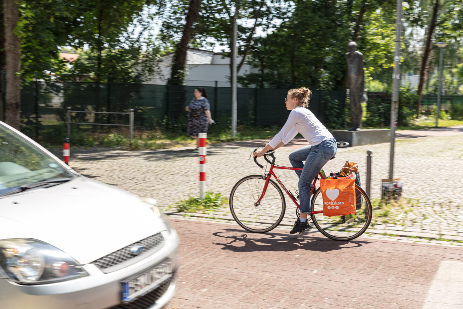 Eine Person fährt Fahrrad vorbei an einem Auto und im Hintergrund ist eine Bronzeplastik zu erahnen.
