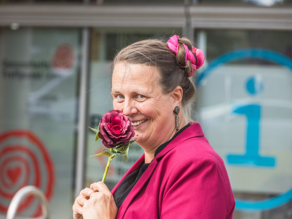 Eine Person hat ihre Haare zu einem Dutt hochgesteckt und trägtdazu Blumen im Haar. Außerdem trägt sie eine Rose in einer Hand die sie an ihr lächelndes Gesicht hält.