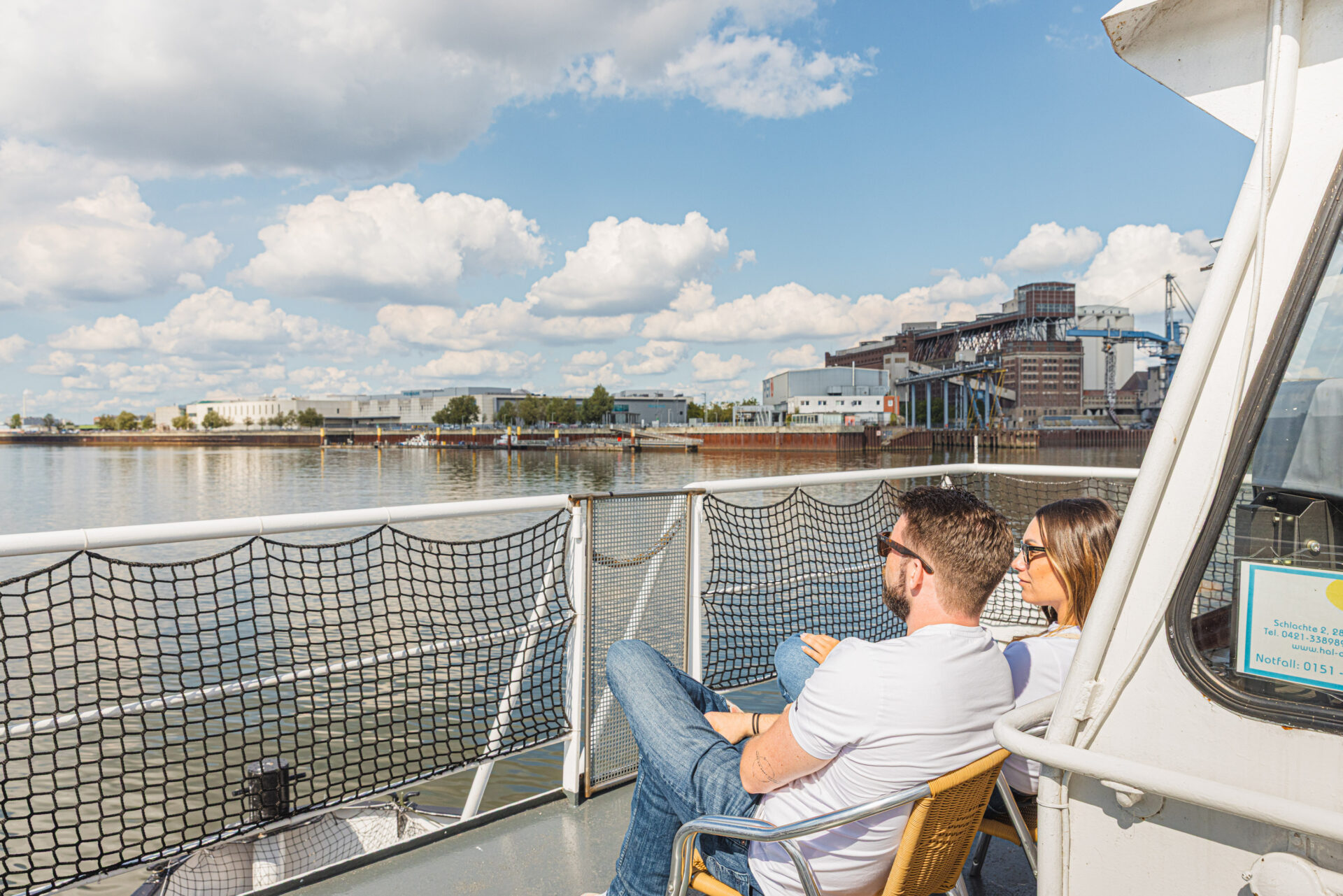 Zwei Personen sitzen auf dem Oberdeck der Weserfähre Bremen und schauen über das Wasser in Richtung Land. Zu sehen ist die riesige Getreideanlage Bremen und die Weserpromenade Gröpelingens mit dem Einkaufszentrum  Waterfront Bremen.