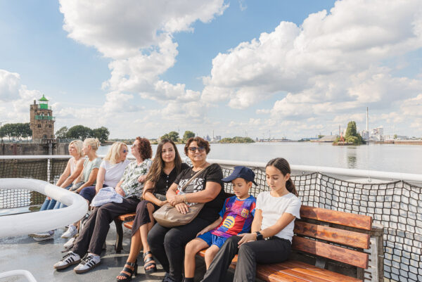 Auf einem Schiff sitzen mehrere Personen auf einer Sitzbank. Im Hintergrund sind Wasser, ein Himmel mit Wolken zu sehen.