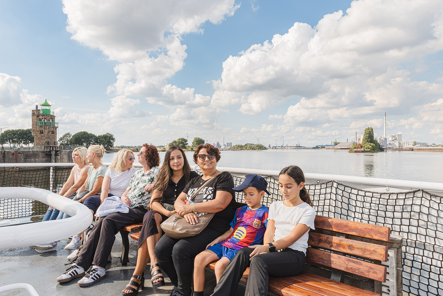 Auf einem Schiff sitzen mehrere Personen auf einer Sitzbank. Im Hintergrund sind Wasser, ein Himmel mit Wolken zu sehen.
