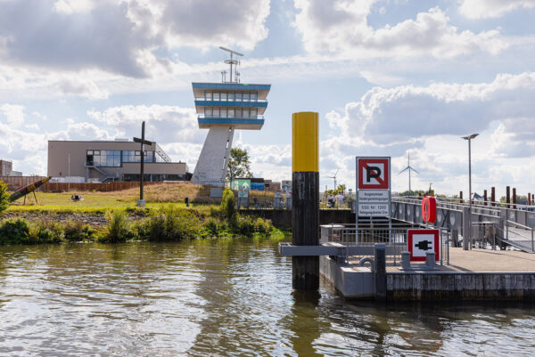 An einem Uferbereich steht ein Turm mit mehreren Ebenen und einer Radaranlage auf der Spitze. Im Vordergrund befinden sich ein Steg mit Schildern, ein Rettungsring und verschiedene Poller. Links steht eine skulpturartige Konstruktion in Raketenform, während auf der Wiese ein kreuzförmiges Metallobjekt zu sehen ist. In der Ferne stehen mehrere Windkraftanlagen vor einem hellen Himmel.