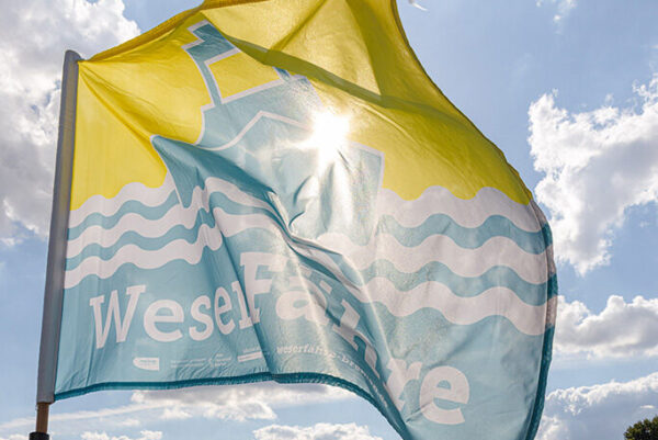 Eine Flagge mit der Aufschrift „WeserFähre“ weht im Wind vor einem Gewässer. Die Flagge zeigt ein blau-gelbes Design mit Wellenmotiven und einem stilisierten grafischen Element. Im Hintergrund sind Kräne und weitere industrielle Strukturen am gegenüberliegenden Ufer zu sehen. Rechts im Bild befindet sich ein kleines Boot auf dem Wasser.