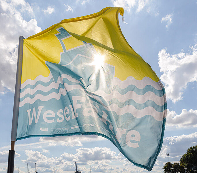 Eine Flagge mit der Aufschrift „WeserFähre“ weht im Wind vor einem Gewässer. Die Flagge zeigt ein blau-gelbes Design mit Wellenmotiven und einem stilisierten grafischen Element. Im Hintergrund sind Kräne und weitere industrielle Strukturen am gegenüberliegenden Ufer zu sehen. Rechts im Bild befindet sich ein kleines Boot auf dem Wasser.