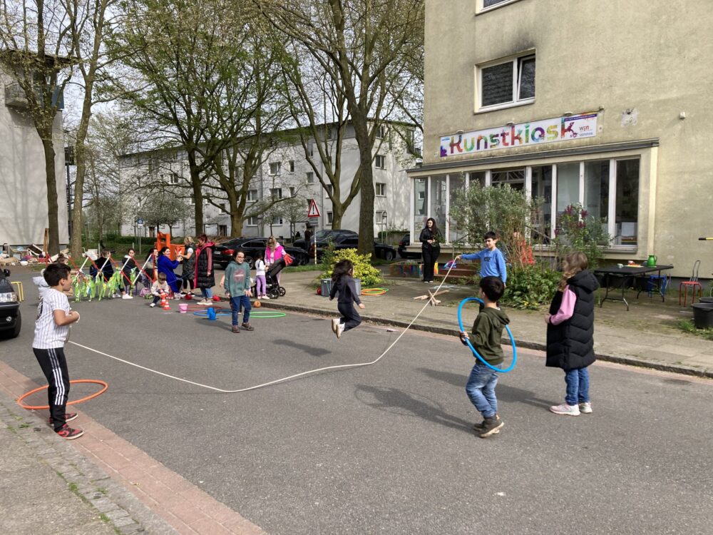 Kinder spielen auf einer für den Verkehr abgesperrten Straße. Sie spielen zum Beispiel Seilhüpfen.