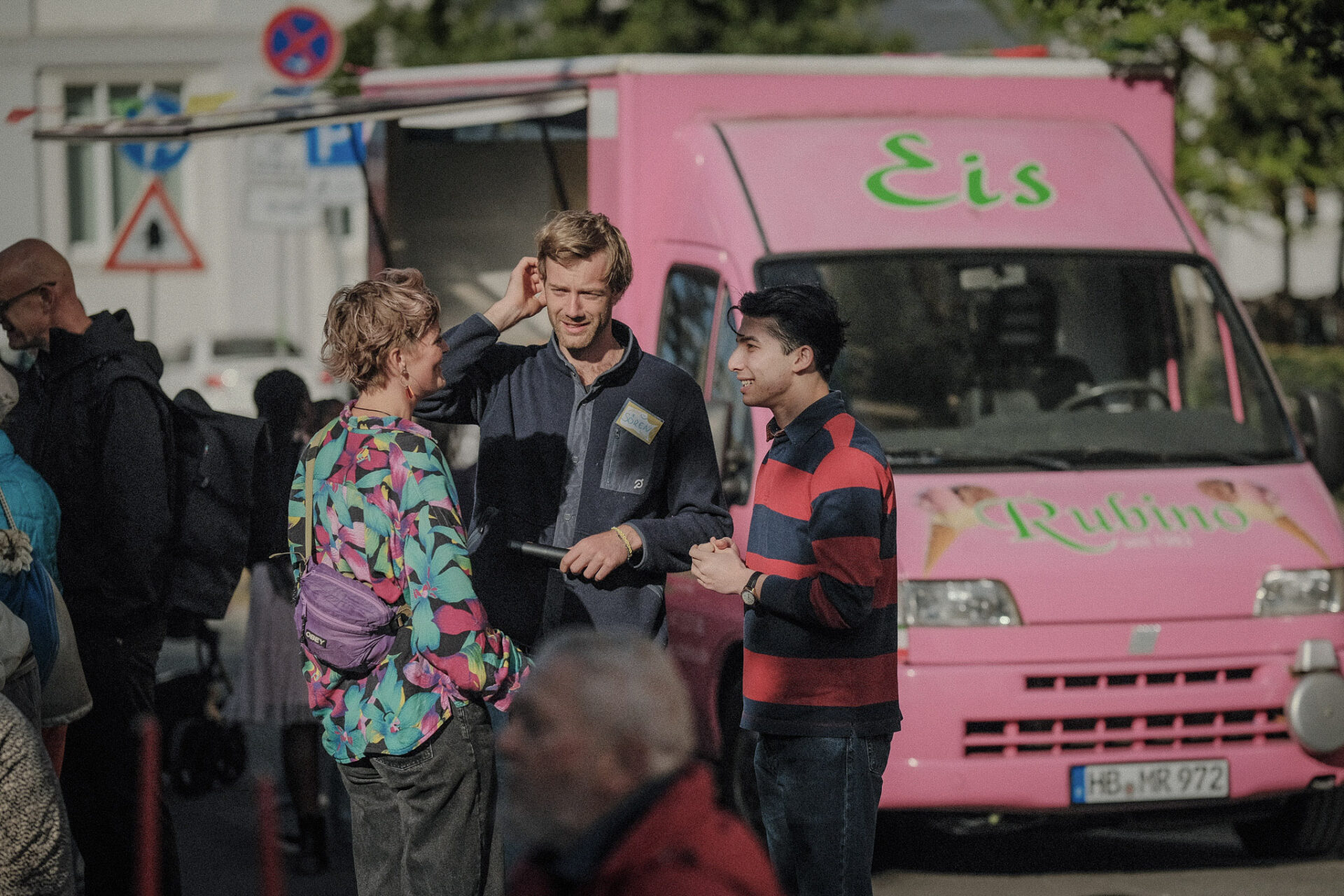 Das Team von Zu Tisch, Svenja, Asad und Sören unterhalten sich auf der Straße. Im Hintergrund steht der Rosa Eiswagen von Eis Rubino.