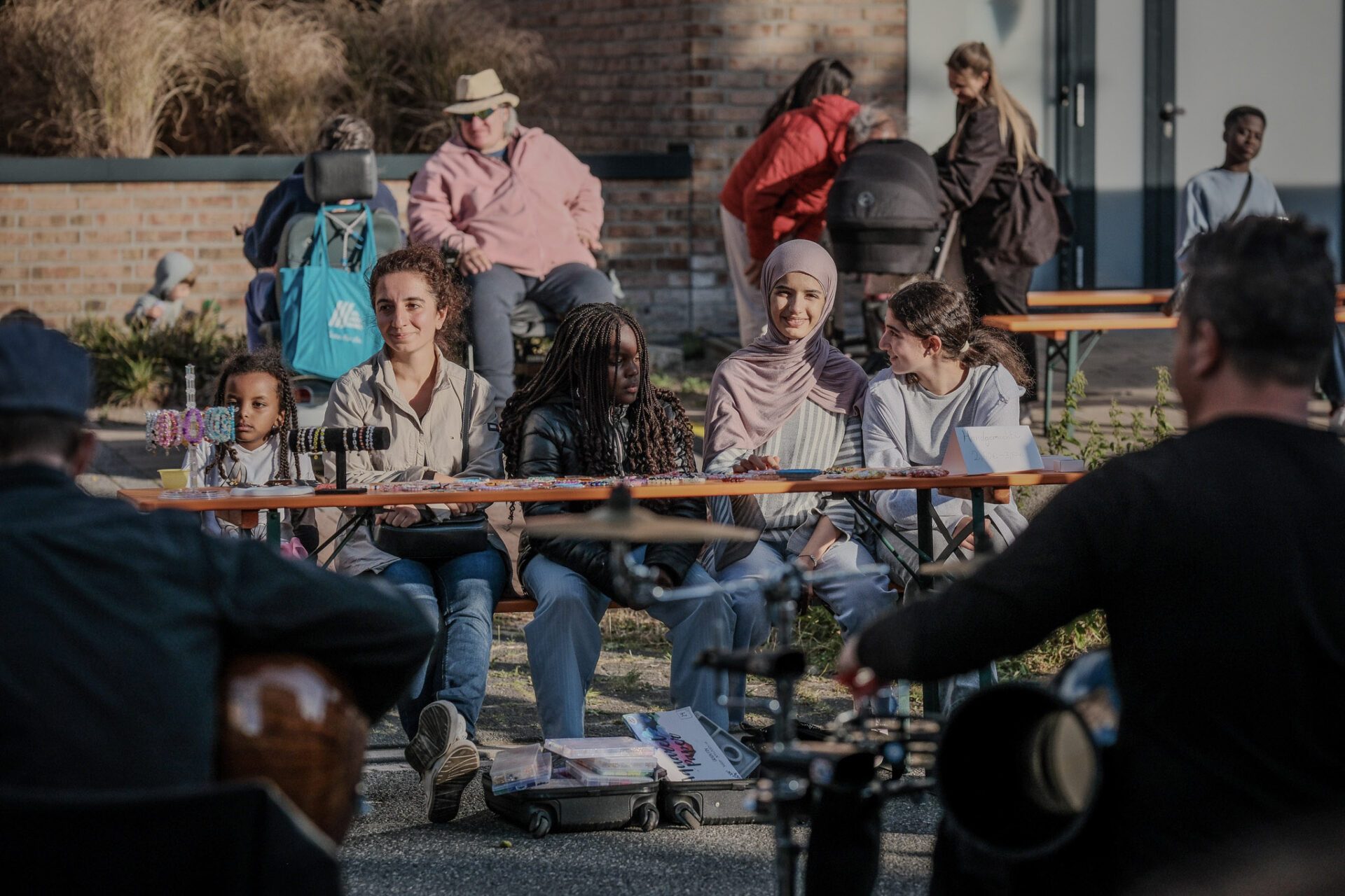 Der Fokus des Bildes liegt auf einem Tisch an dem vier Kinder und eine Frau sitzen. Sie machen Armbänder an ihrem Tisch.