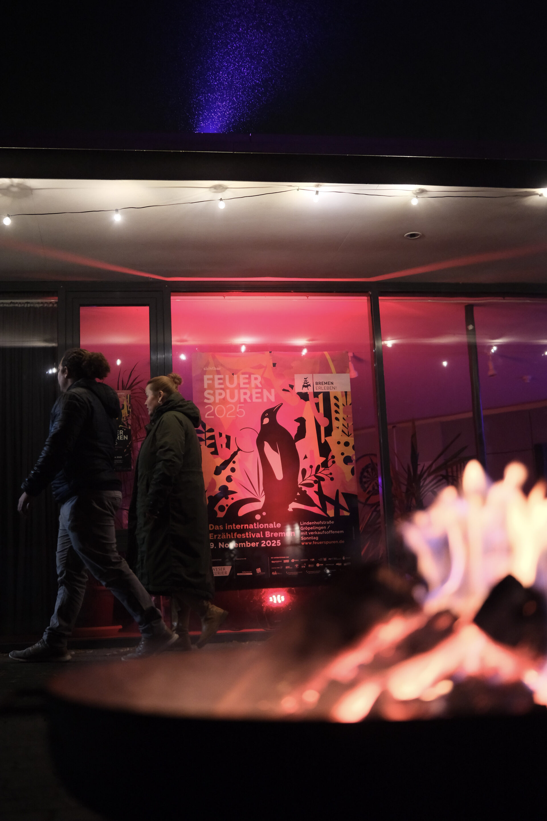 Zwei Personen gehen an einer rot beleuchteten Fensterfront vorbei in der ein großes Plakat mit einem Pinguin drauf zu sehen ist.
