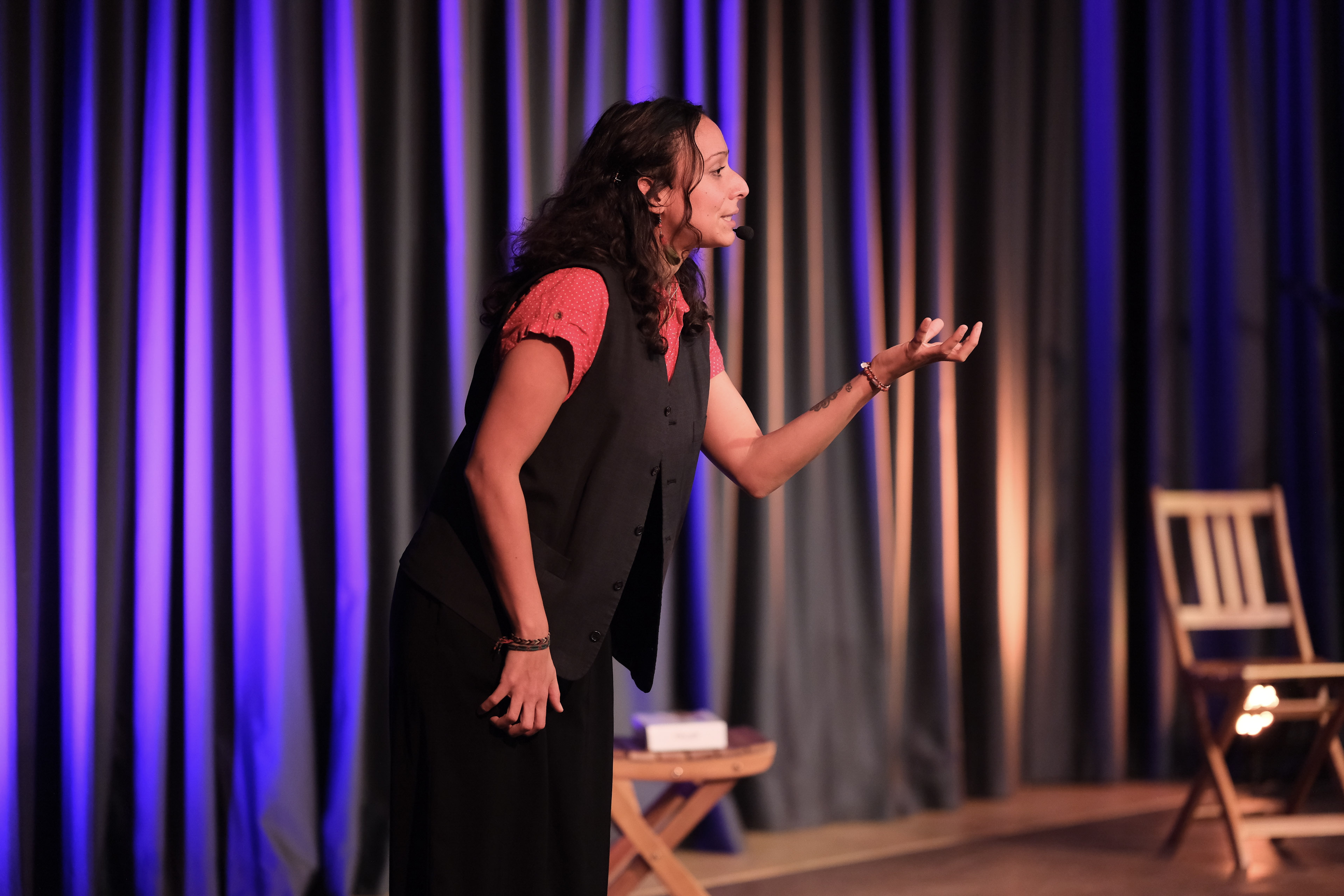 Vor einem Bühnenvorhand gestikuliert eine Person auf der Bühne mit den Armen. Im Hintergrund sind zwei Holzstühle zu sehen.