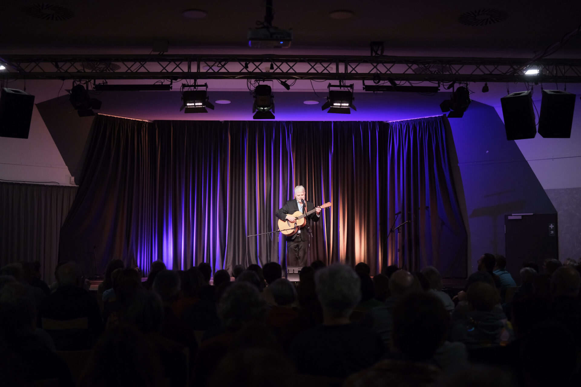 Eine Person sitzt auf einer Bühne mit der Gitarre in der Hand und spielt vor einem großen Publikum, das nur in Andeutung zu sehen ist im abgedunkelten Saal.