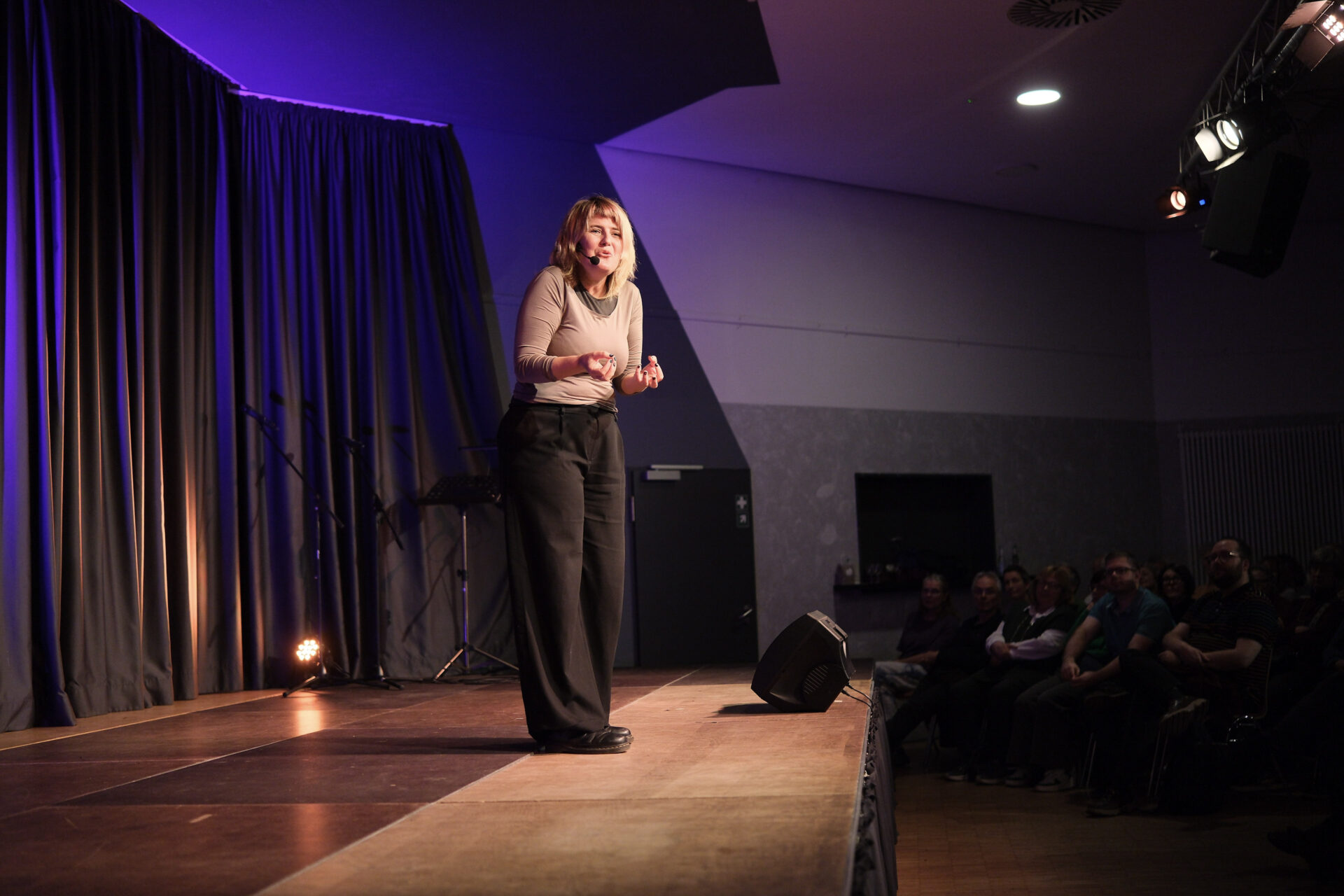 Auf einer Bühne steht im eine Person, die in unsere Richtung schaut, spricht und dabei ausdruckstark gestikuliert.