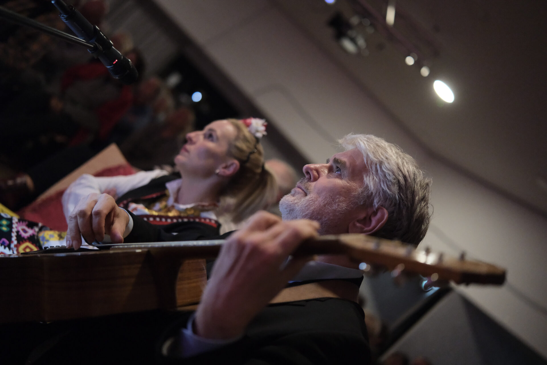 Eine Person in folkloristischer Tracht mit Blumenkranz im Haar und eine Person mit einer Gitarre schauen rechts nach links.