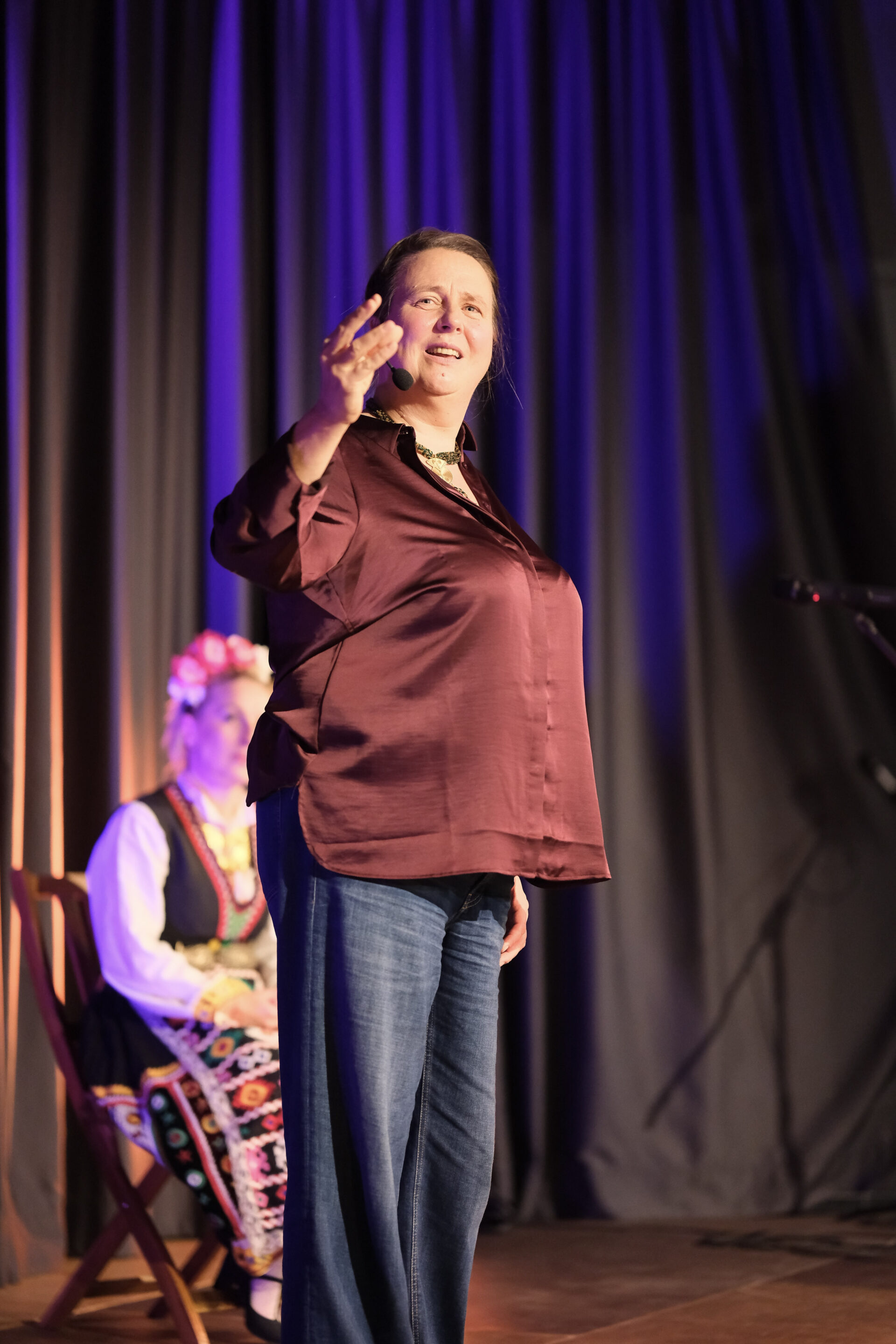 Auf einer Bühne steht im Vordergrund eine Person, die in unsere Richtung schaut und dabei ihre rechte Hand in ausdruckstarker Geste hochhält. Hinter ihr sitzt eine weitere Person in folkloristischer Tracht.