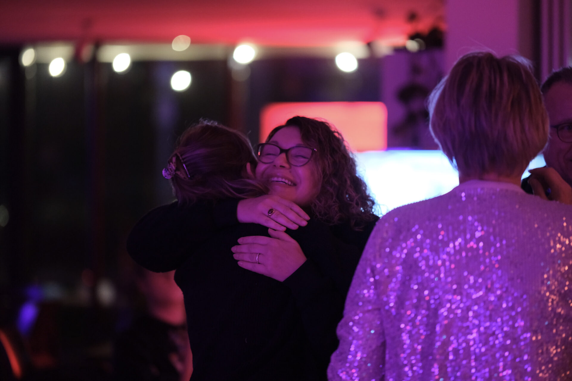 In einem festlich beleucteten Raum umarmen sich zwei Personen herzlich. Weitere Personen sind schemenhaft zu sehen.