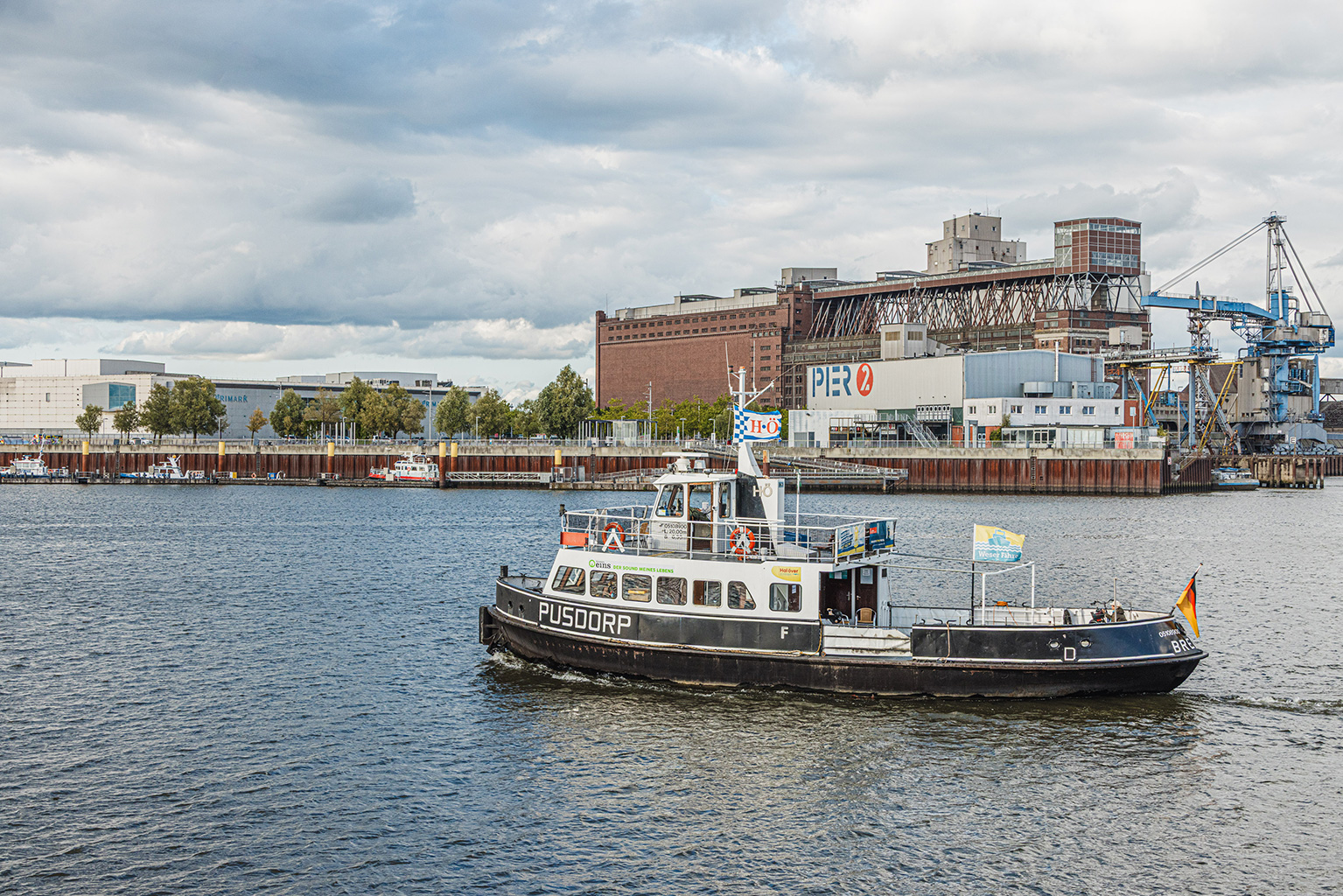 Auf einem Gewässer fährt eine kleine Personenfähre mit dem Namen „PUSDORP“. Das Schiff ist schwarz‑weiß lackiert und trägt mehrere Flaggen, darunter eine am Heck. Im Hintergrund stehen Industriegebäude, Kräne und ein großes Gebäude mit der Aufschrift „PIER 2“ unter einem teils bewölkten Himmel.