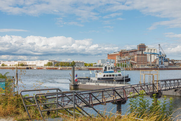 An einer Uferböschung befindet sich der Steg zu einem Schwimmanleger. An dem Anleger liegt ein kleines Fahrgastschiff. Im Hintergrund ist ein Hafenbecken sowie ein gigantisches Industrie.gebäude zu sehen