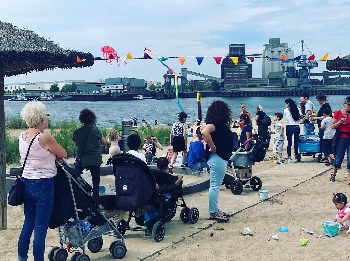 Ein belebter Strandabschnitt mit vielen Erwachsenen und Kindern. Kinderwagen, Sandspielzeug und bunte Wimpel prägen das Bild. Im Hintergrund fliegt ein Drachen am Himmel, dahinter liegt ein Industriehafen am Wasser.