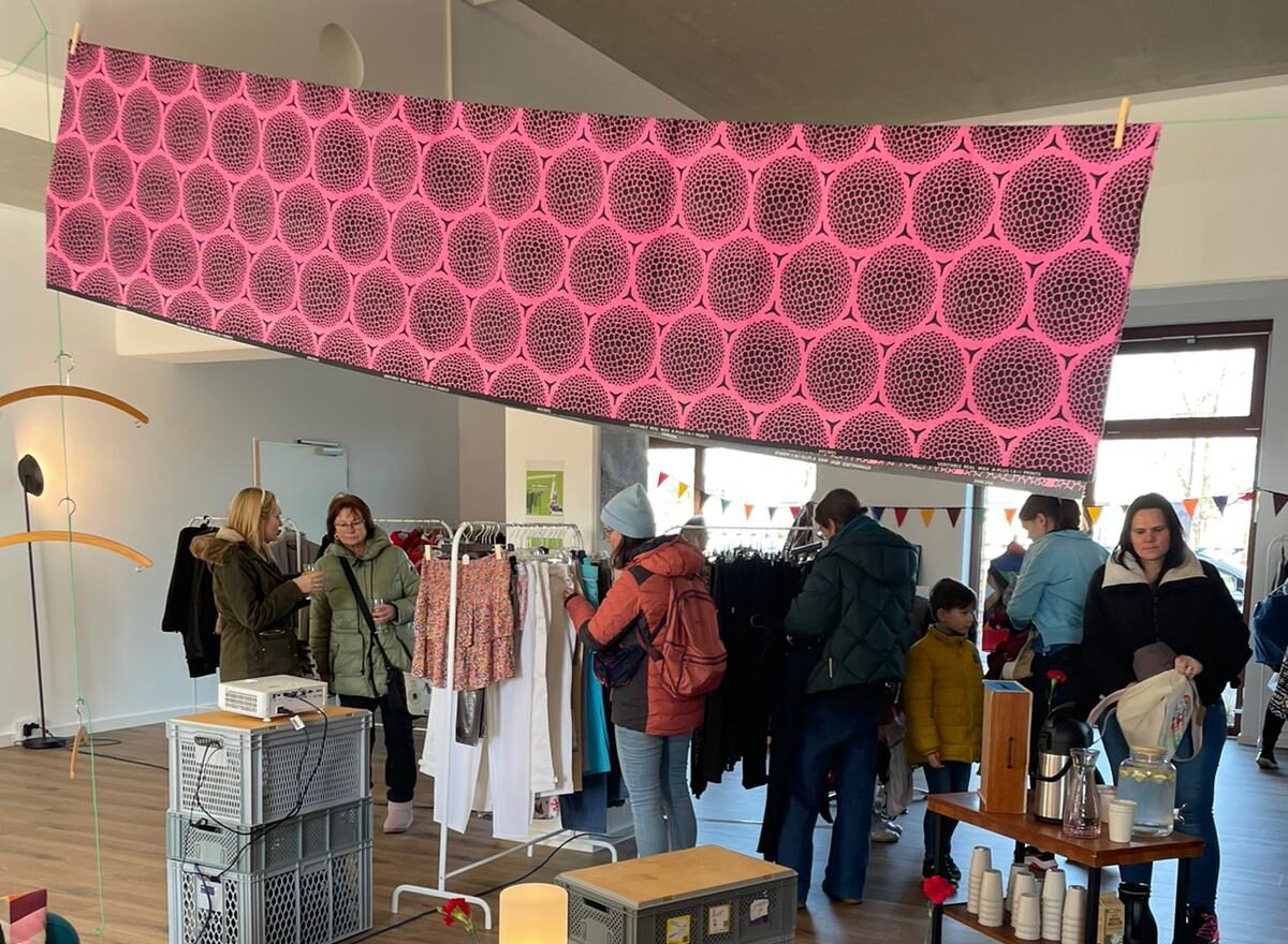 In einem Raum hängt unter der Decke ein langes buntes Tuch. Darunter sind Menschen, die an Kleiderständern die Klamotten begutachten. Auf dem Boden stehen Sitzmöbel aus Europaletten und Eurokisten.