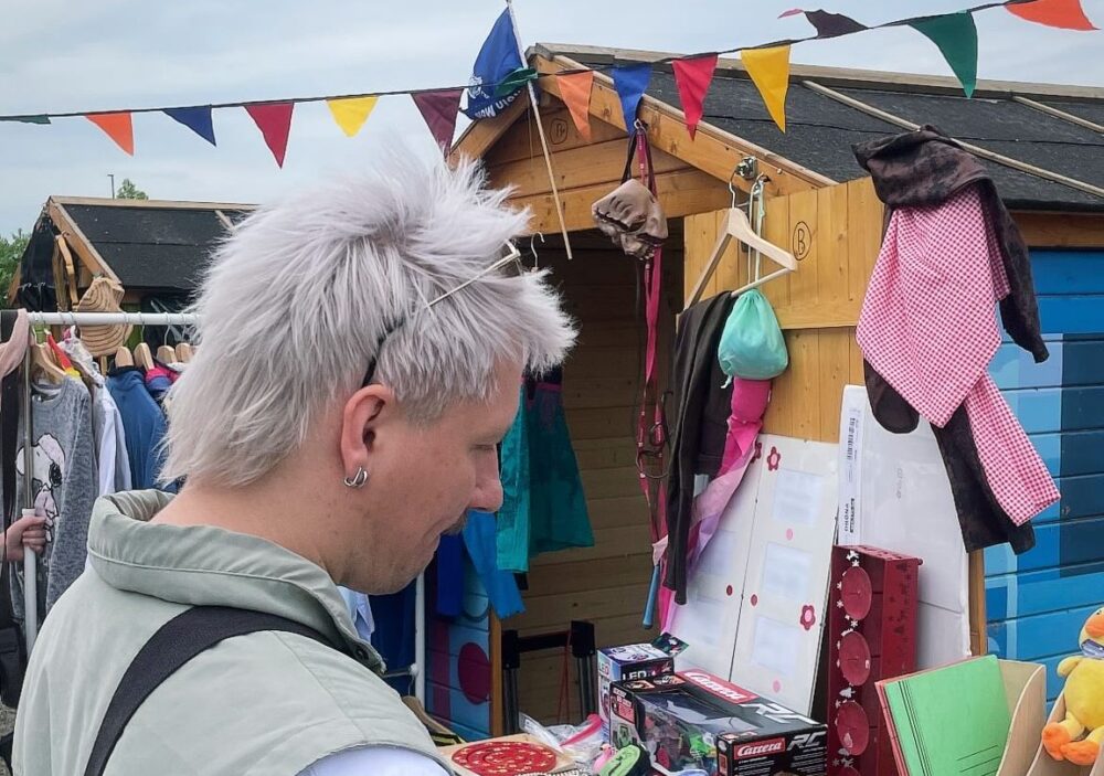 Eine Person steht vor einer Holzbude und einem Flohmarkstand und guckst sich die Sachen an. Eine bunte Wimpelkette flattert im Wind.