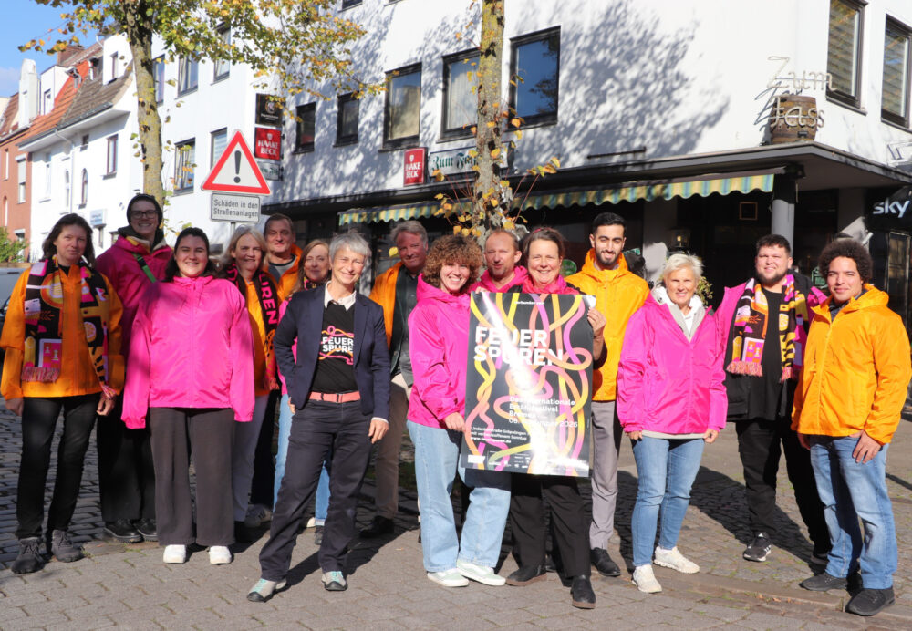 Eine Gruppe von Menschen steht auf der Straße, einige von Ihnen tragen pinke oder orangene Jacken. Es wird auch ein Plakat hochgehalten mit der Werbung für das Erzählfestival Feuerspuren 2026