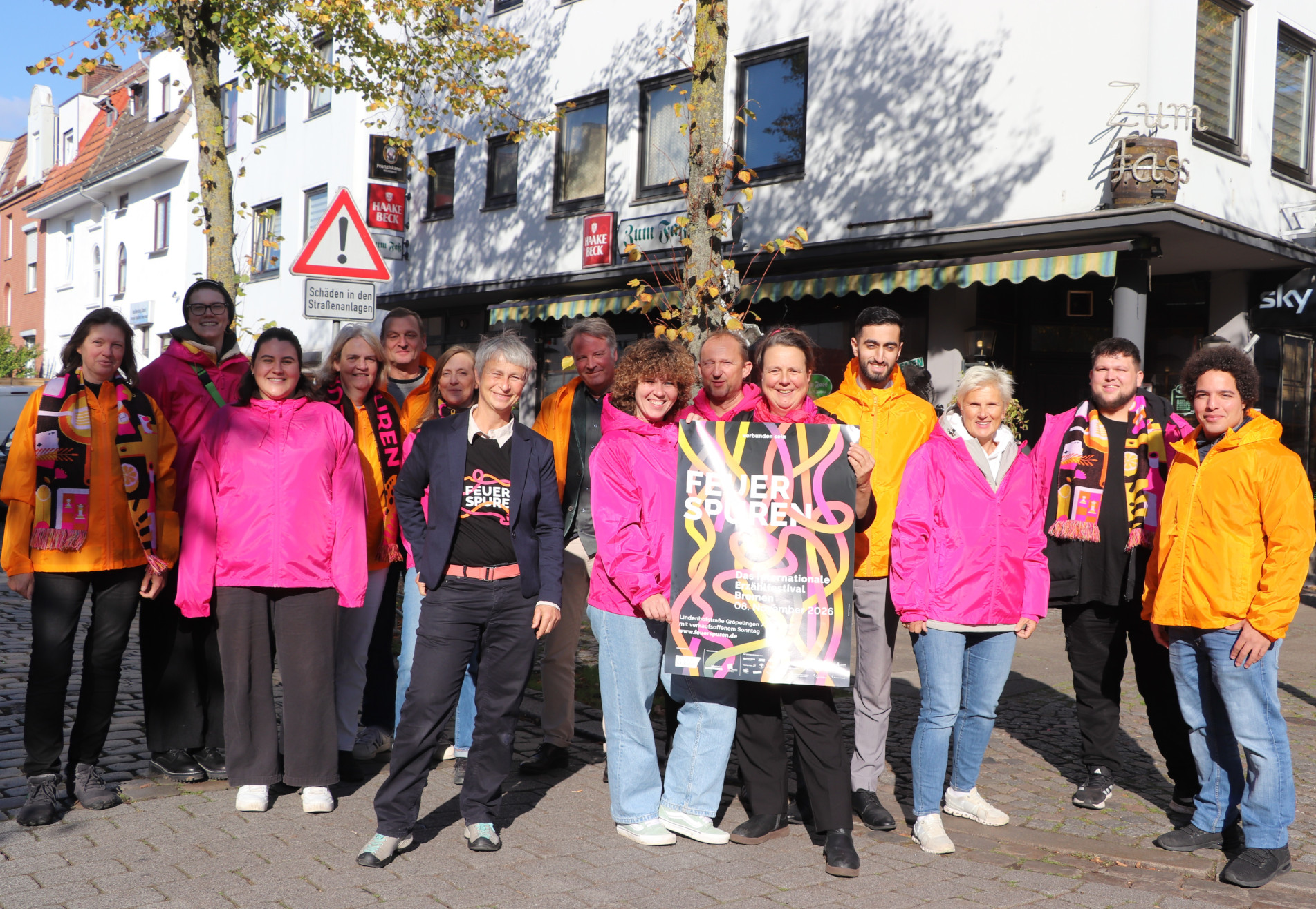 Eine Gruppe von Menschen steht auf der Straße, einige von Ihnen tragen pinke oder orangene Jacken. Es wird auch ein Plakat hochgehalten mit der Werbung für das Erzählfestival Feuerspuren 2026