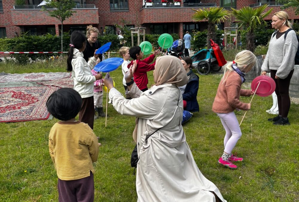 12. Mehrere Kinder und Erwachsene spielen auf einer Wiese vor einem mehrstöckigen Backsteingebäude mit bunten Tellern auf Stäben