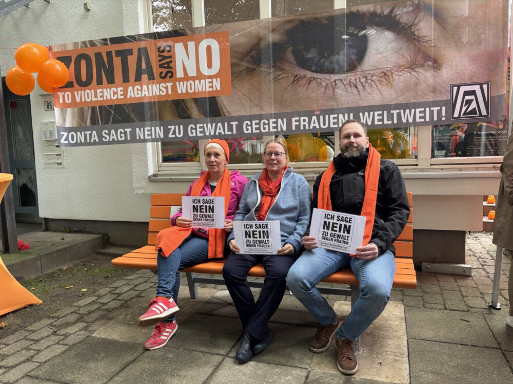 Drei Personen sitzen auf einer orangefarbenen Bank vor einem Gebäude. Hinter ihnen hängt ein großes Banner mit der Aufschrift „Zonta says NO to violence against women“ und „Zonta sagt Nein zu Gewalt gegen Frauen weltweit“. Alle drei halten Schilder mit der Aufschrift „Ich sage Nein zu Gewalt gegen Frauen“ und tragen orangefarbene Schals.